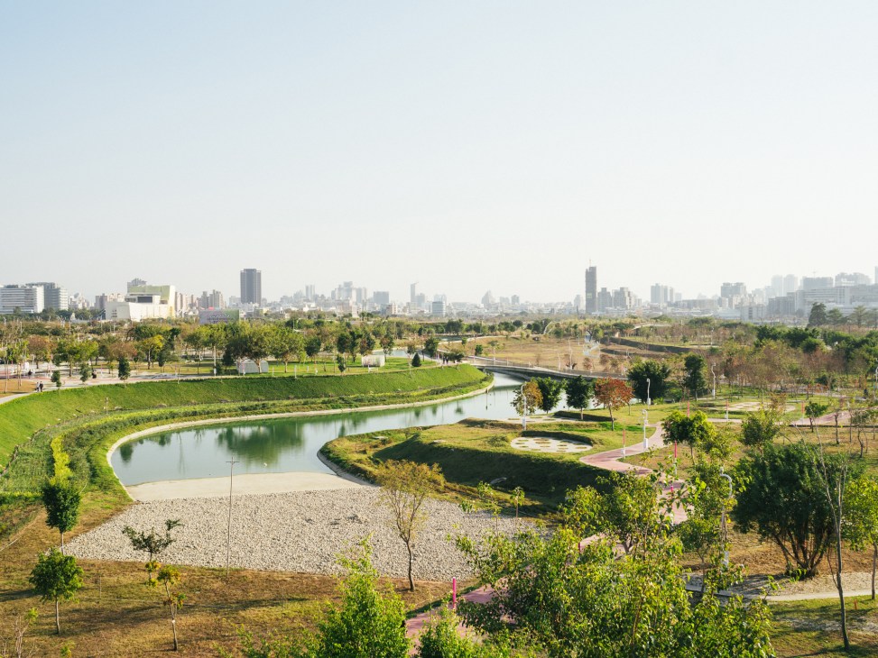 A winding waterway cuts through landscaped parkland with walking paths, planted areas, and a city skyline in the background