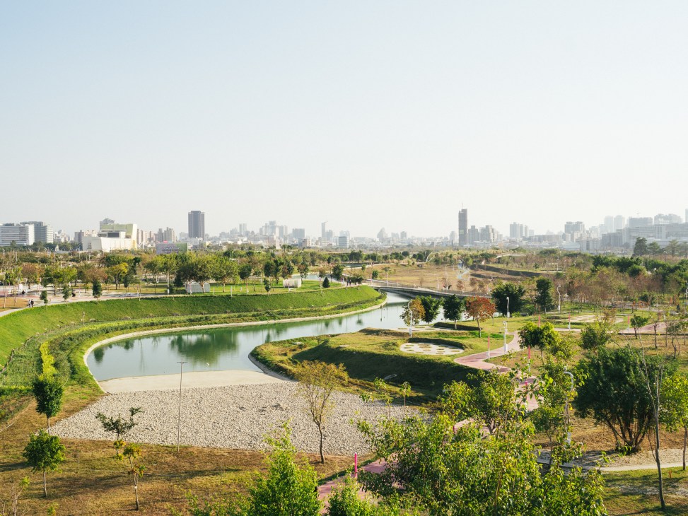Modern urban park with curved water feature, green spaces, and city skyline in background