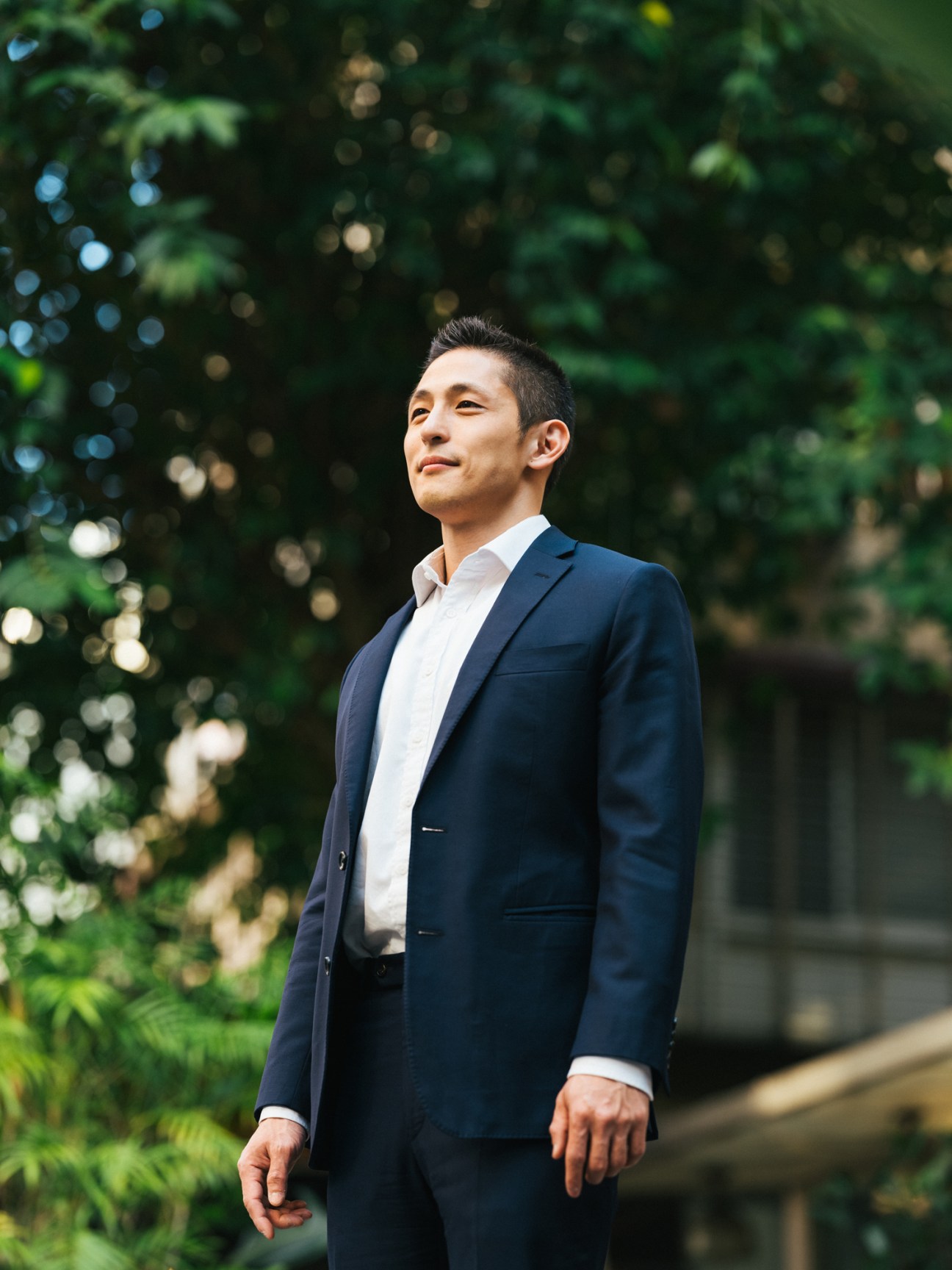 Enoch Wu in a navy suit jacket and white shirt looking upward outdoors with green foliage in background