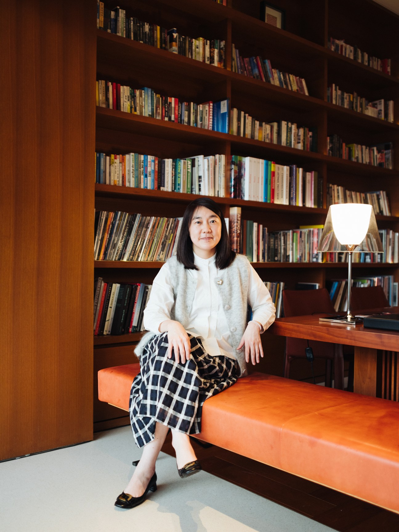 Woman in cream cardigan and checkered skirt sitting on orange bench in library with floor-to-ceiling bookshelves