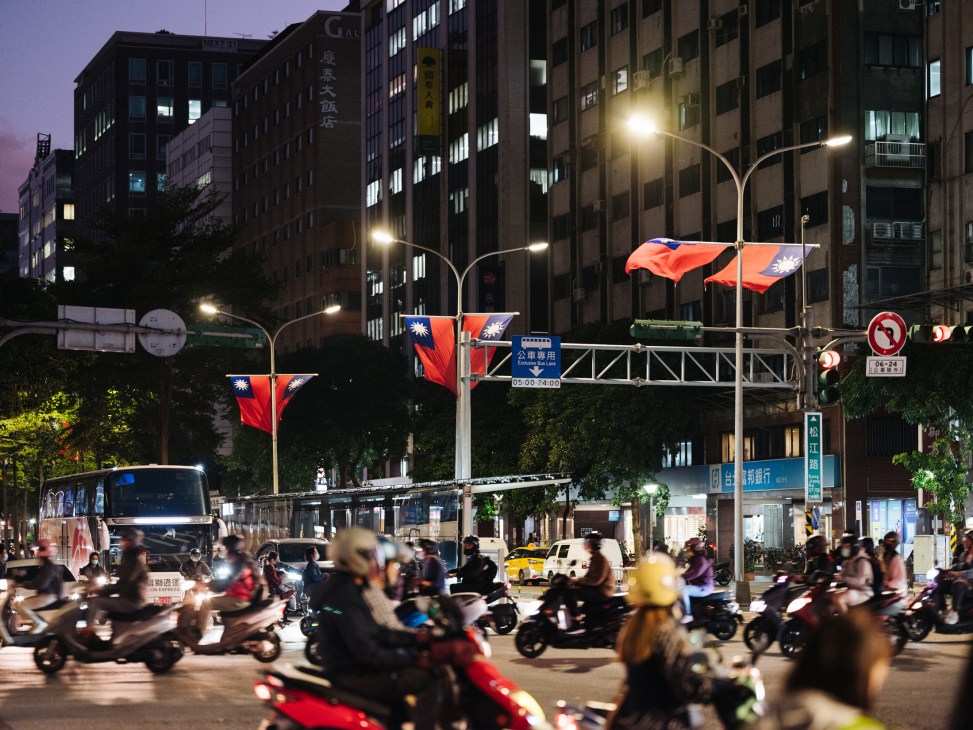 Busy downtown Taipei street at dusk with Taiwanese flags hanging from streetlights and scooters in motion.