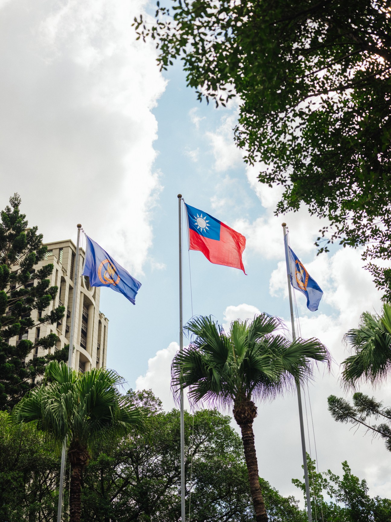 Taiwan's national flag flying on a flagpole among palm trees in Taipei with blue sky and clouds