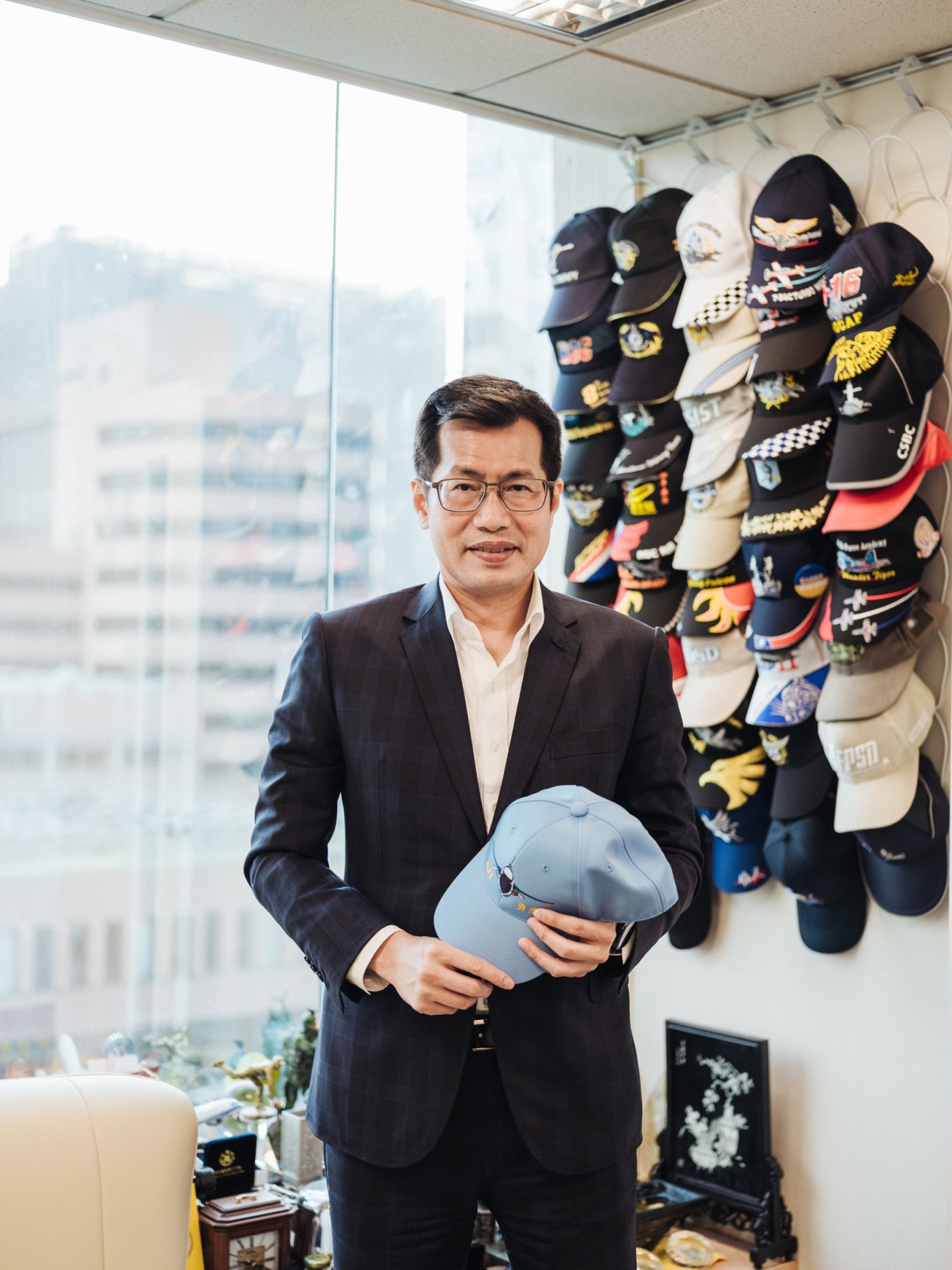 Lo Chih-cheng, a Democratic Progressive Party MP, poses in an office holding a light blue cap with decorative masks displayed on the wall behind him.