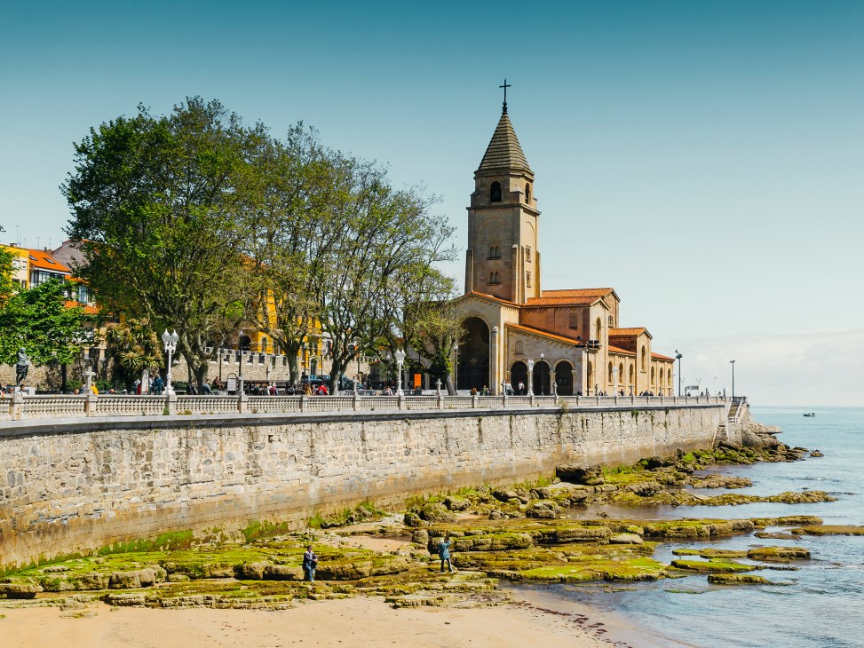 Coastal promenade in Gijón with a church, yellow buildings and sandy beach along the waterfront