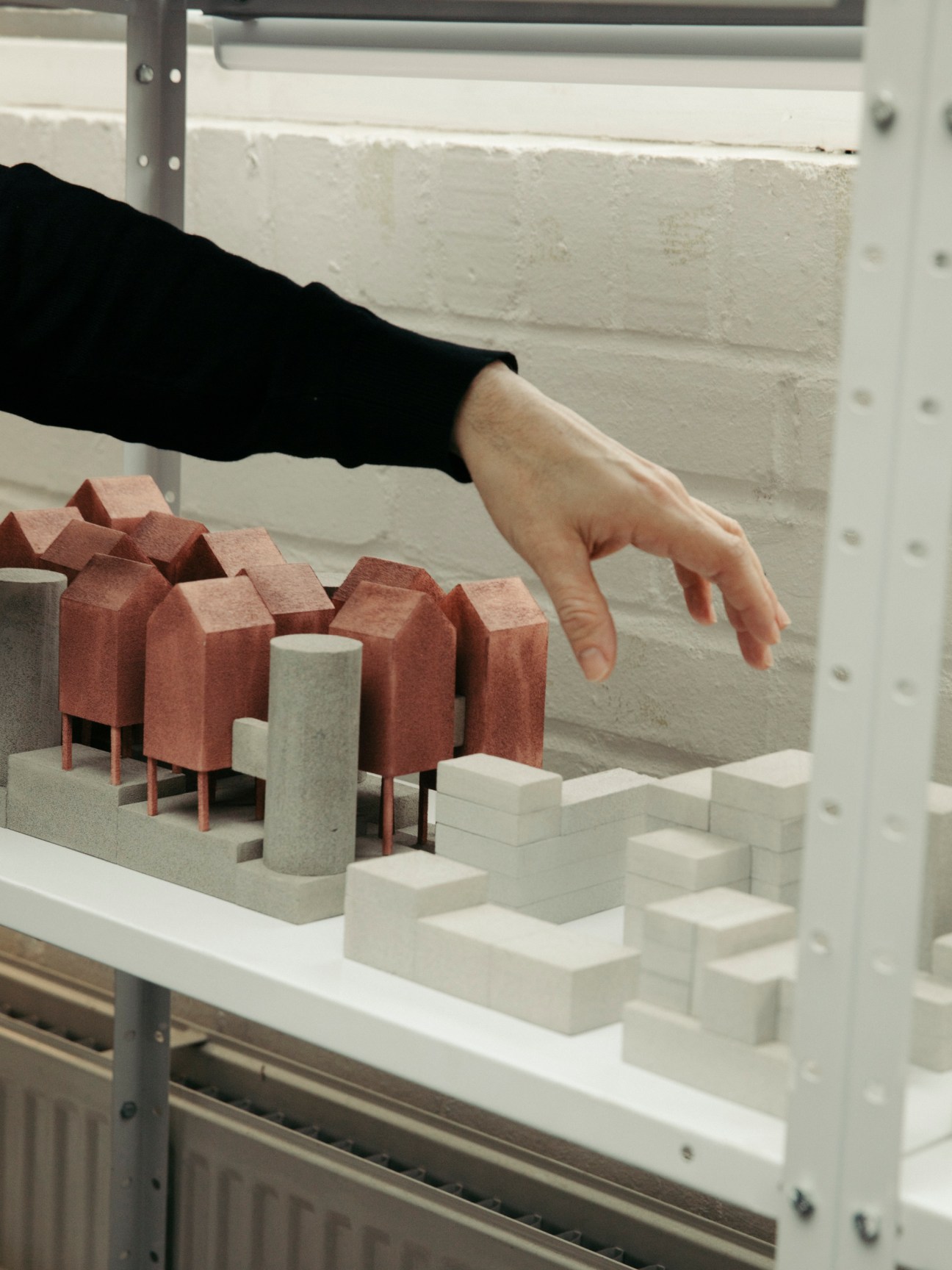 Hand reaching toward architectural model with red and white miniature building components on white surface