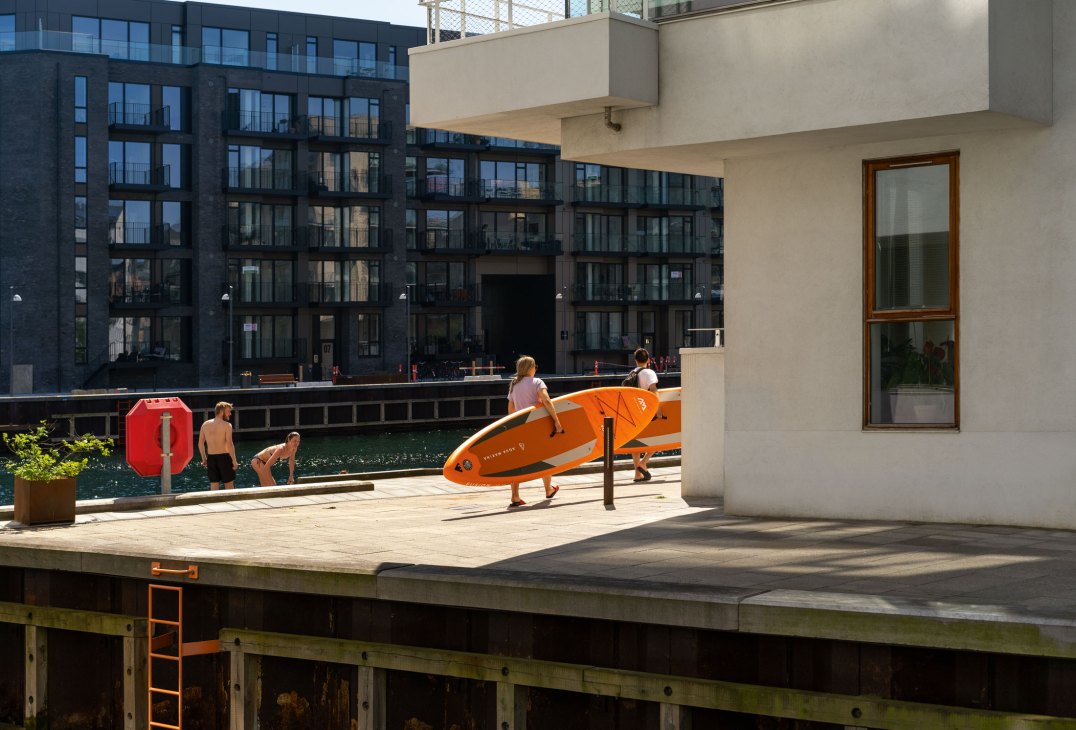People with an orange surfboard by a waterfront promenade with modern buildings in Copenhagen