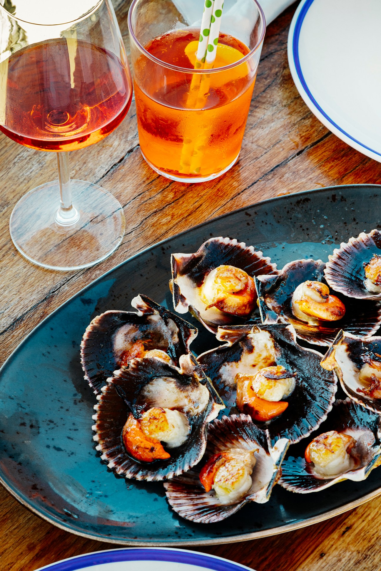 Scallop shells on a dark plate with amber cocktails and wine on a wooden table.