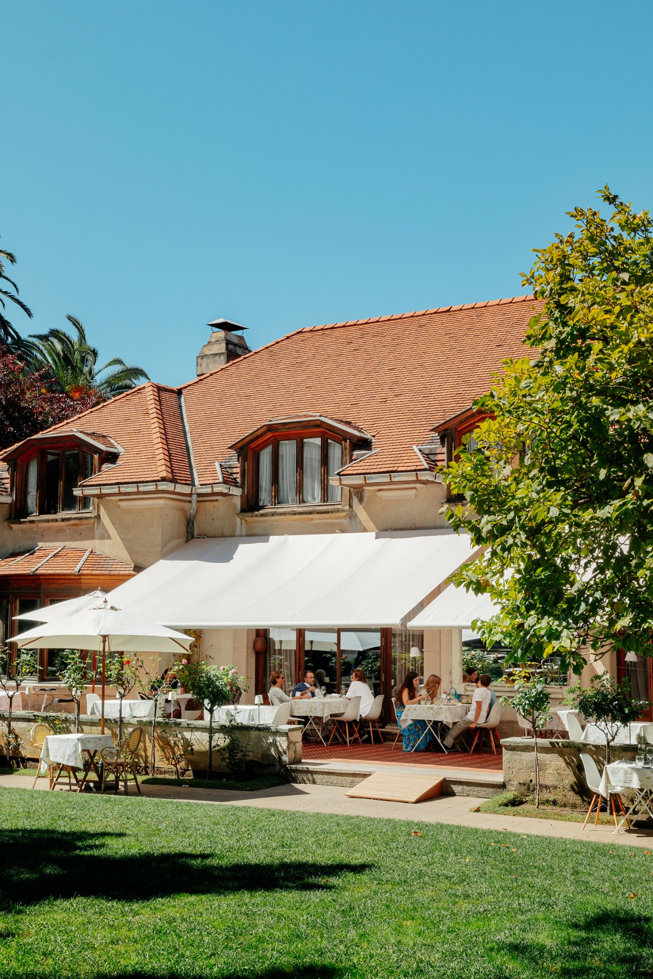 Outdoor restaurant terrace with white umbrellas and diners seated at tables beside a building with orange tile roof