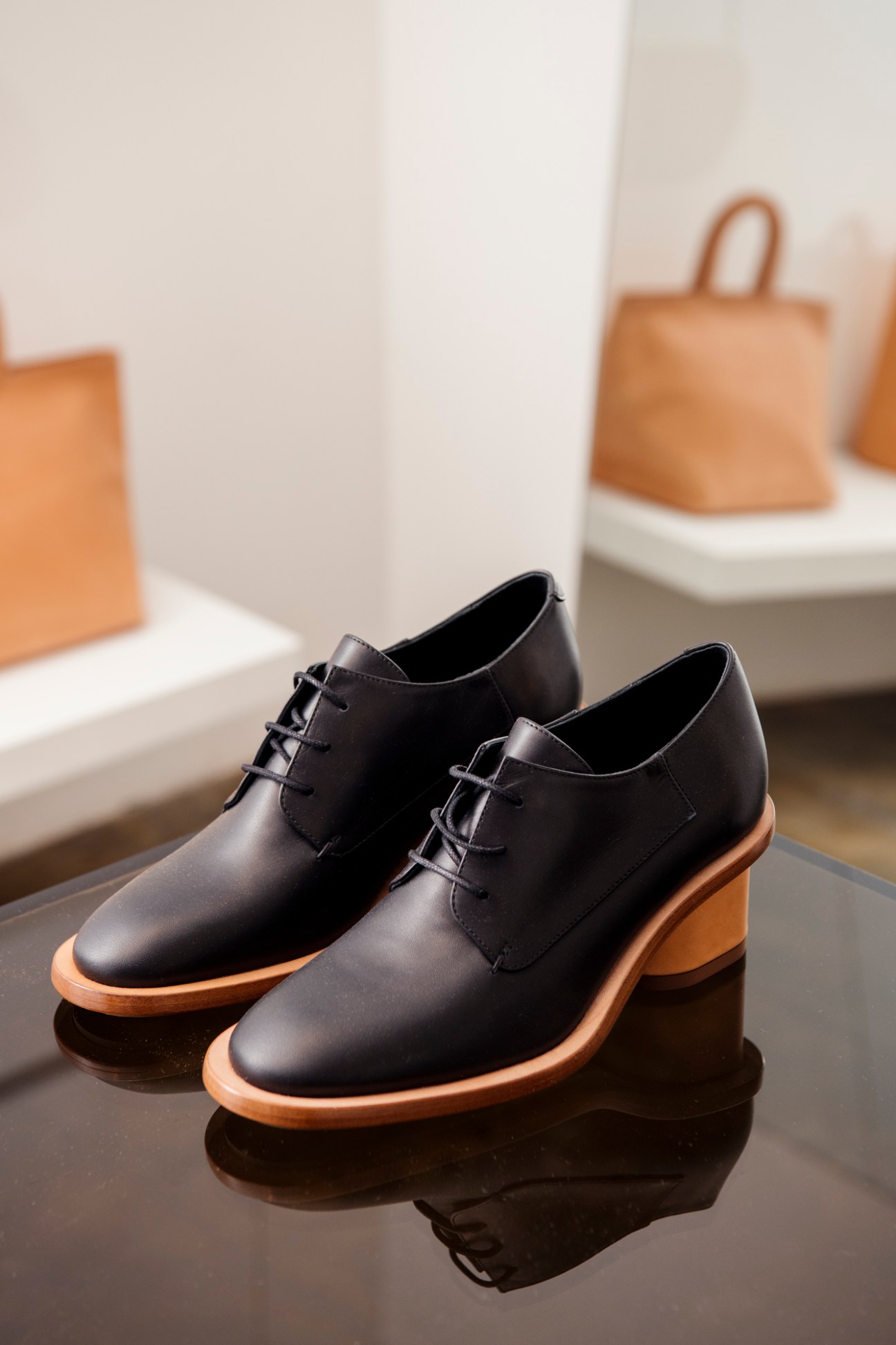 Black leather oxford shoes with natural wood soles displayed on a dark table with tan leather bags blurred in background.