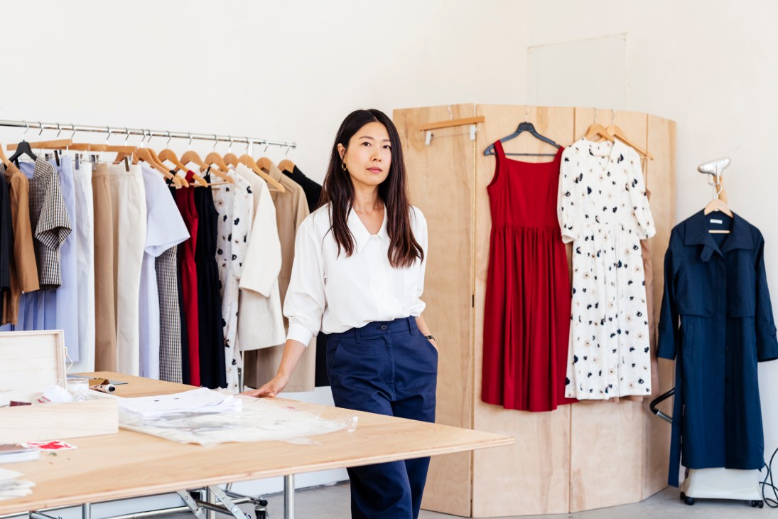 Jeana Sohn stands in her studio with clothing designs on hangers and work materials on a table.