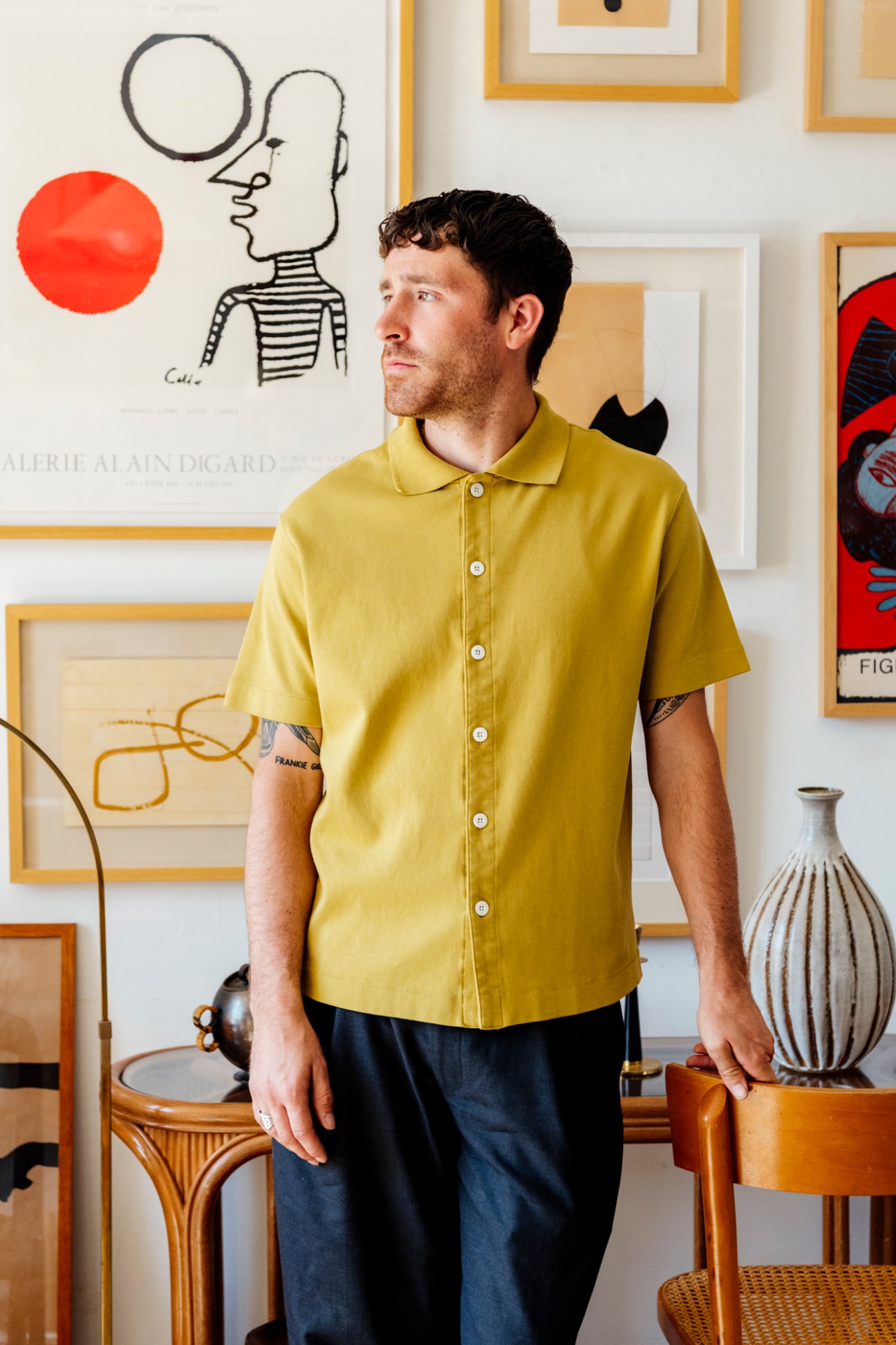 Man in yellow button-up shirt standing in front of framed artworks and a ceramic vase.