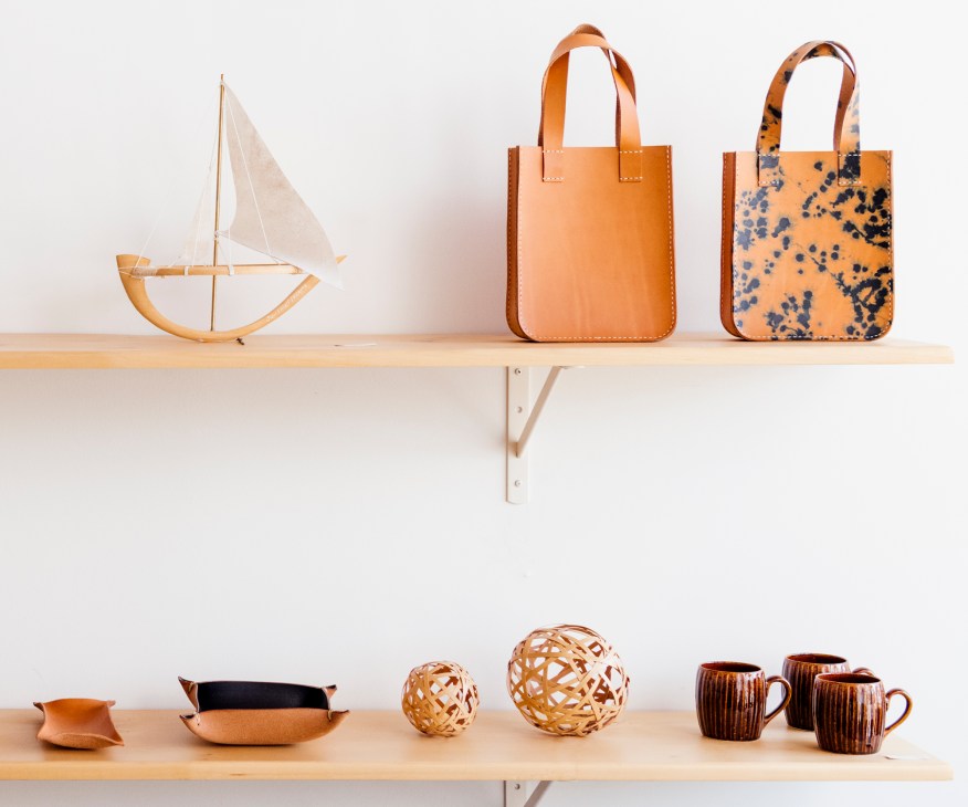Wooden shelf displaying Made Solid leather goods including a sailboat model, tan totes, woven balls, and ceramic mugs.