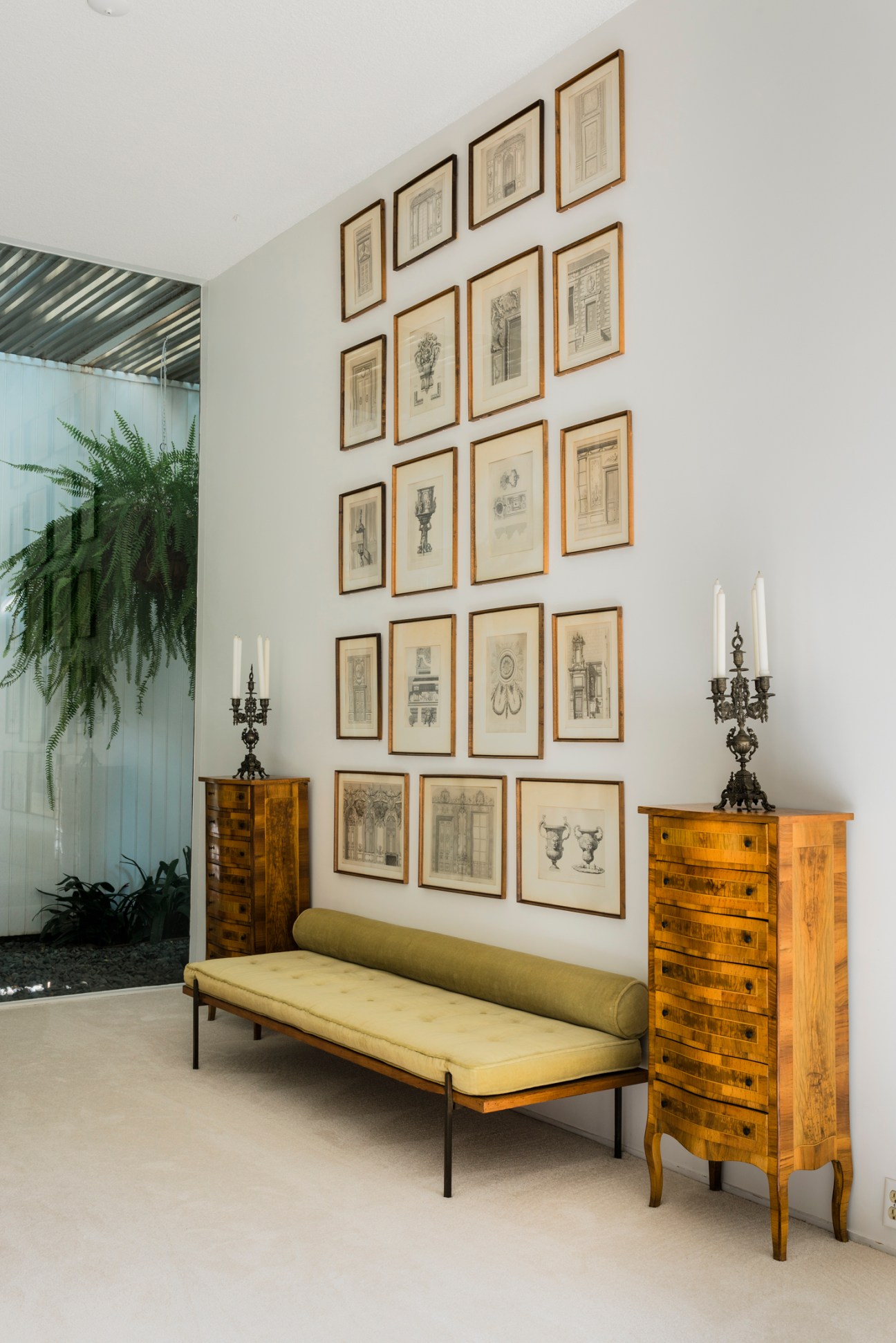 Master bedroom with framed architectural drawings on white wall, yellow bench, and wooden dressers with candelabras.