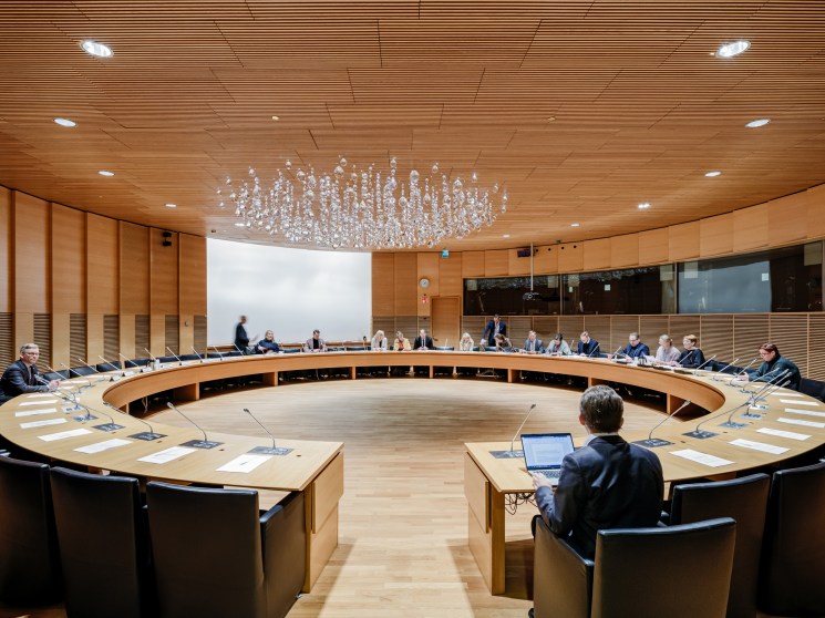 Circular meeting room with wood ceiling and walls, officials seated around curved table with laptops and microphones