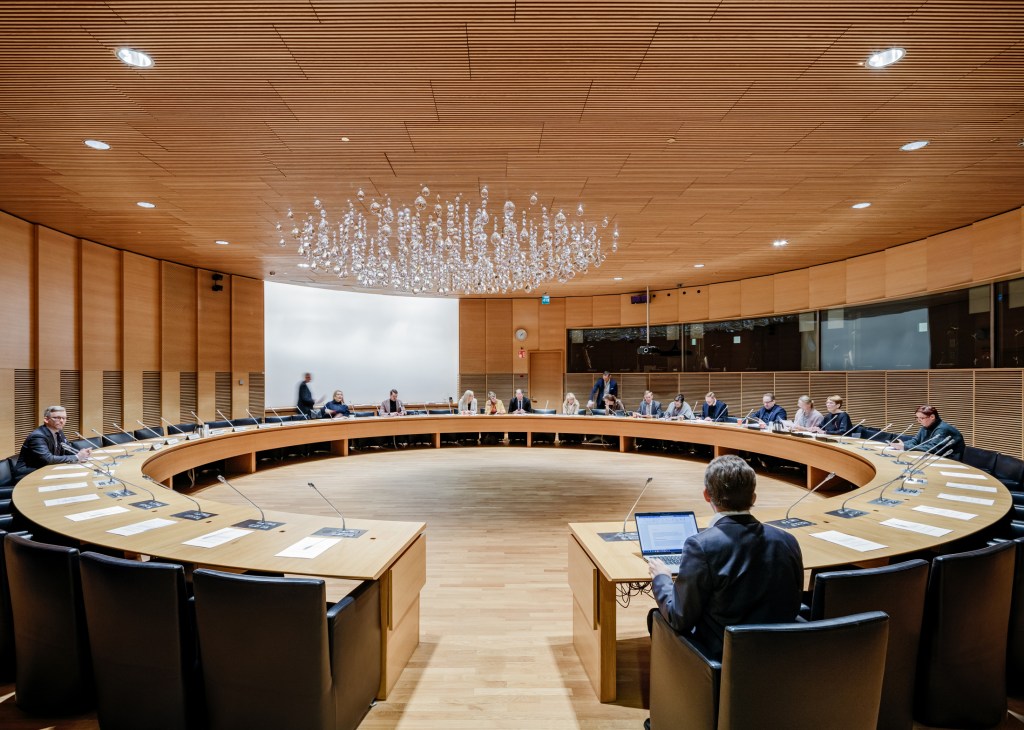 Circular meeting room with wood ceiling and walls, officials seated around curved table with laptops and microphones