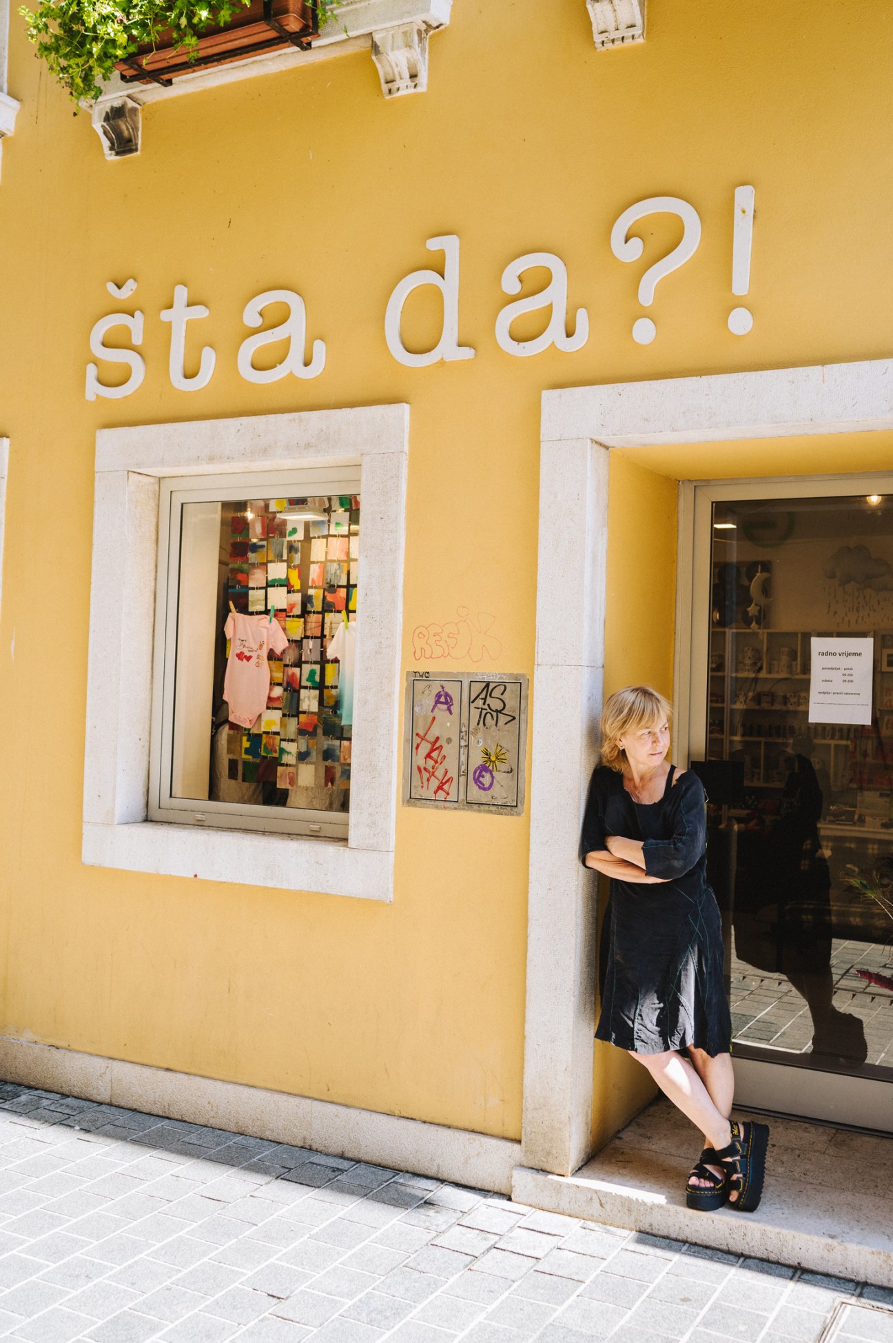 Woman in dark dress standing in doorway of yellow building with white text 'šta da?!' above storefront