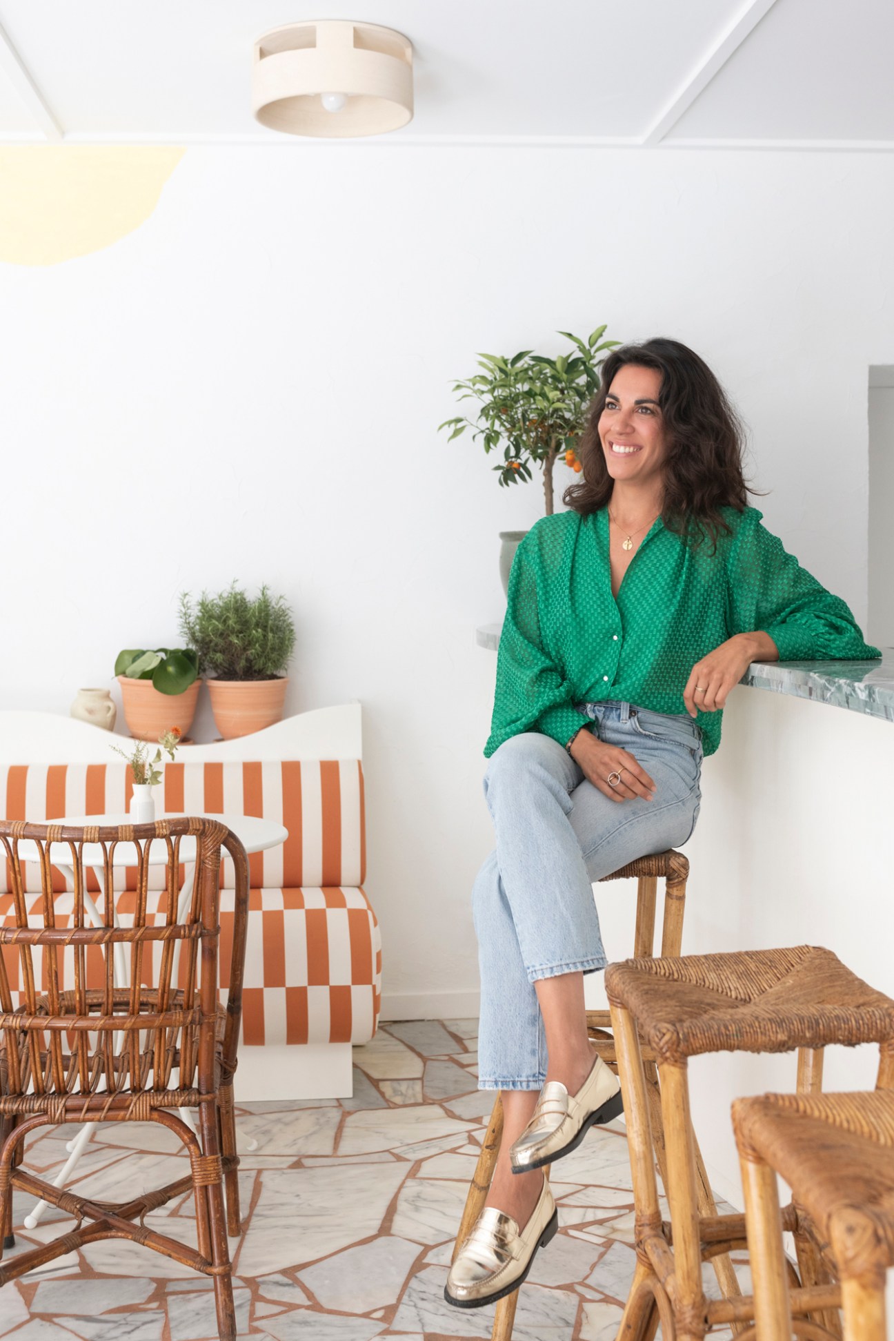 Woman in green shirt sitting on wicker stool in bright Mediterranean-style interior with terracotta tiled floor