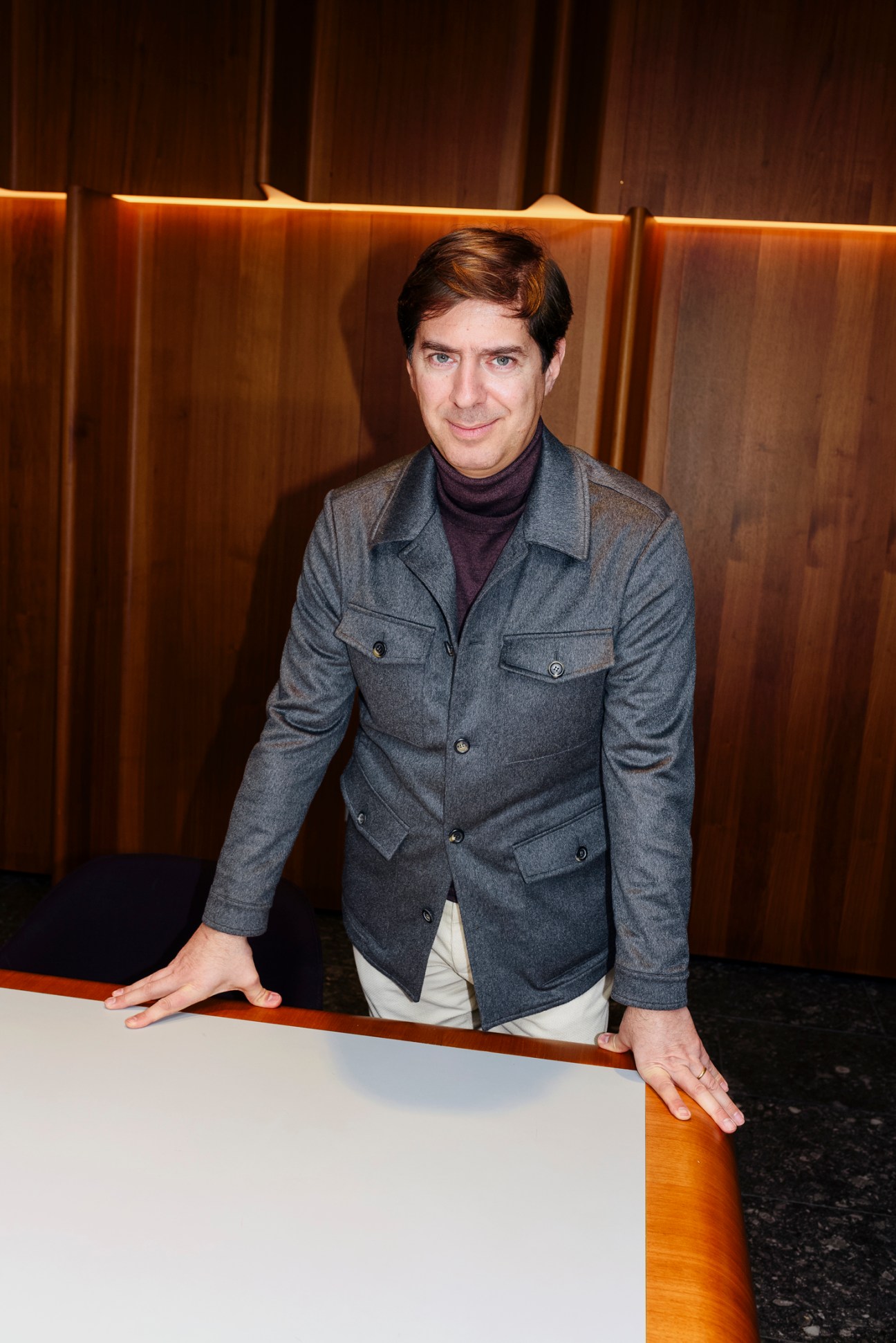 Stefano Canali leaning against a wooden counter in a modern interior wearing a grey jacket and dark turtleneck