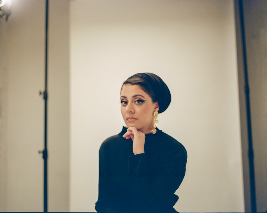 Sumayya Vally in a black turtleneck sweater with gold earrings, hand positioned thoughtfully under her chin