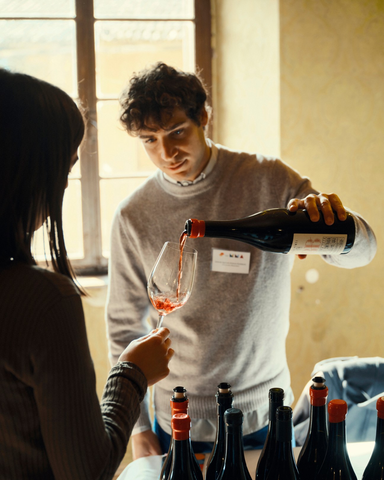 Man in gray sweater pouring red wine from bottle into glass at tasting table with multiple wine bottles