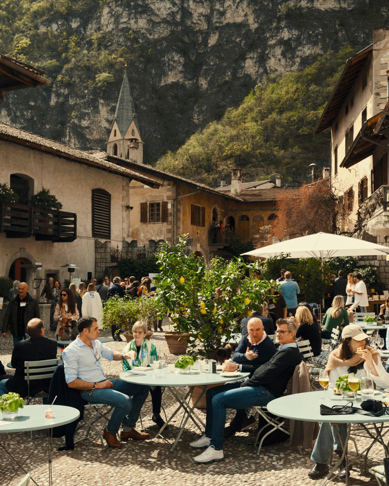People socializing at outdoor cafe tables in a cobblestone courtyard surrounded by historic buildings and mountains