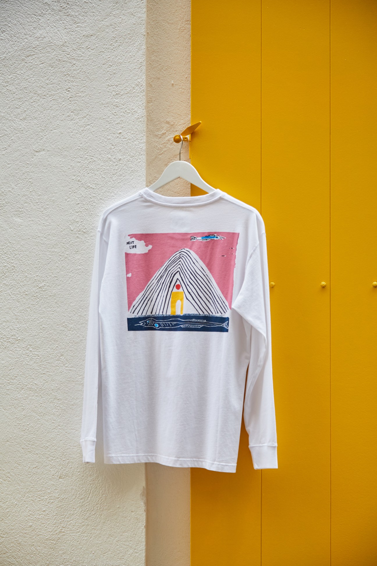 White long-sleeved t-shirt with colorful graphic design hanging on white hanger against yellow wall