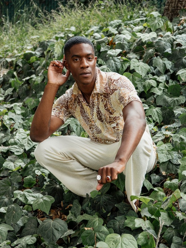 Man wearing patterned shirt sitting cross-legged among green leafy plants