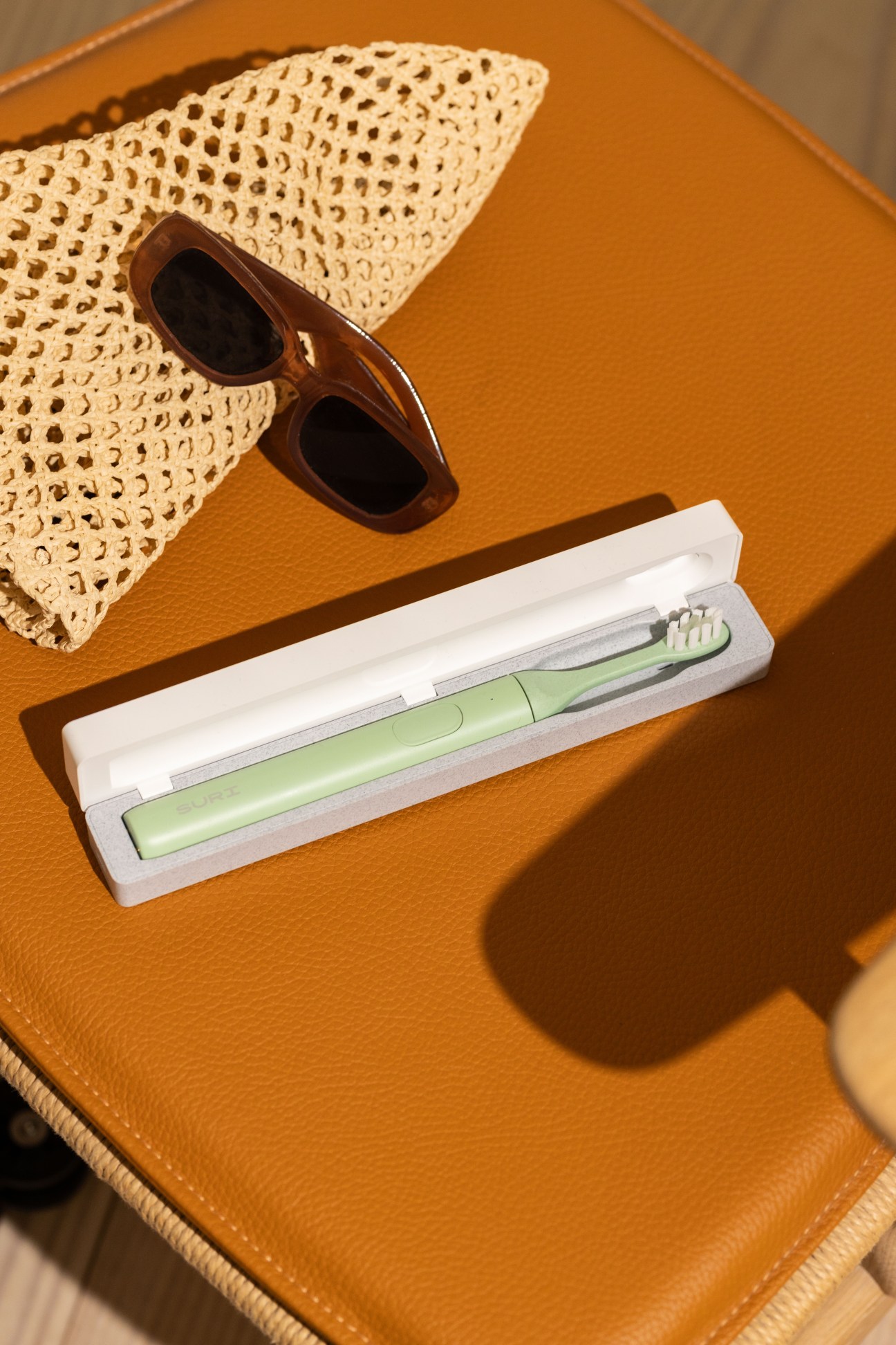 Suri electric toothbrush with plant-based head in mint green displayed in white box with sunglasses and woven hat on orange background.