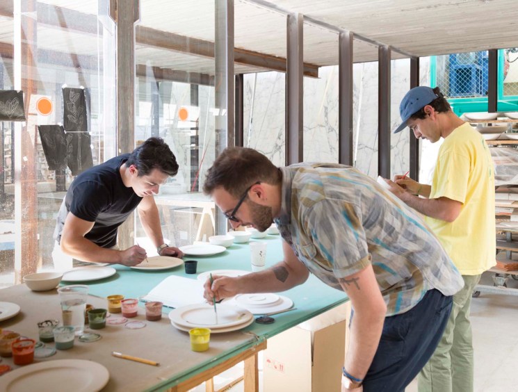 Artists decorating ceramic plates at a factory workshop table during Estación Material art fair.