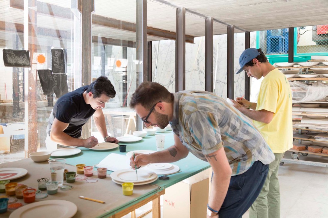 Artists decorating ceramic plates at a factory workshop table during Estación Material art fair.