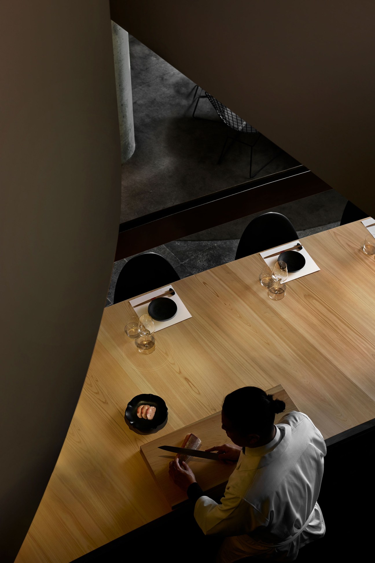 Chef preparing sushi at wooden counter with black plates and tea glasses in dimly lit restaurant