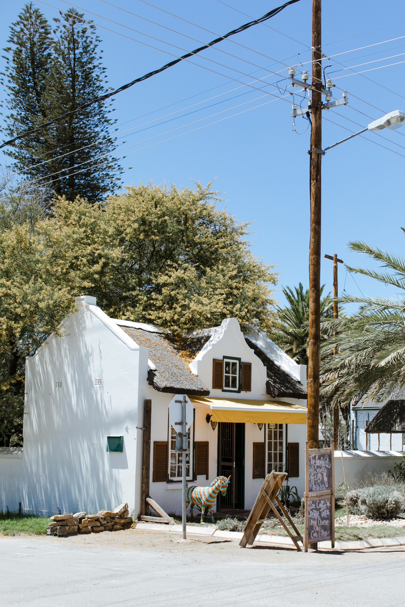 White Cape Dutch-style building with brown shutters, yellow awning, and zebra sculpture outside Die Klein Padstal coffee shop