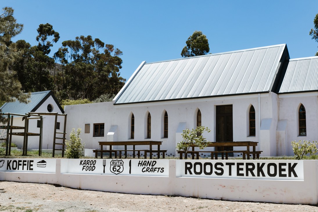 White building with corrugated metal roof and arched windows, featuring signs for coffee, Karoo food, and handcrafts