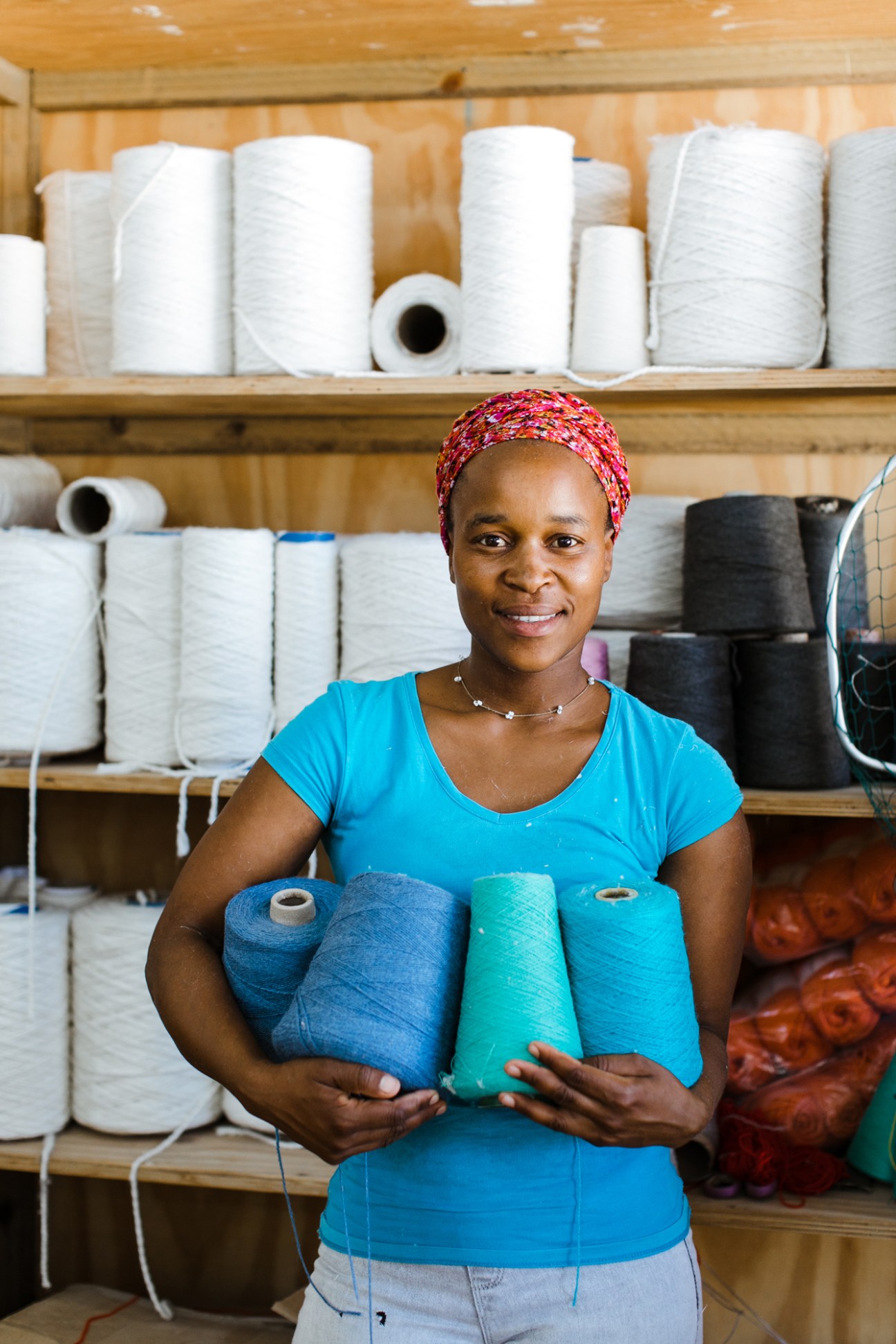 Smiling woman in blue top holding colorful spools of thread at textile workshop with shelves of yarn behind her