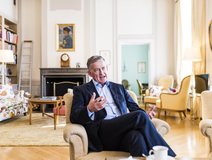 An older man in a suit gestures while speaking from an armchair in an elegant, art-filled sitting room with a fireplace and bookshelves.