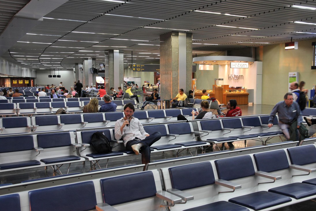 Passengers wait in a sparse airport terminal with rows of blue seating and a departure board visible.