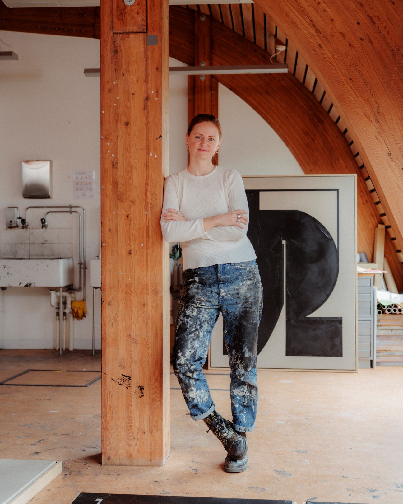 Artist Tabitha Millett standing in her studio with paint-splashed floors and wooden beams, next to a geometric black and white artwork.