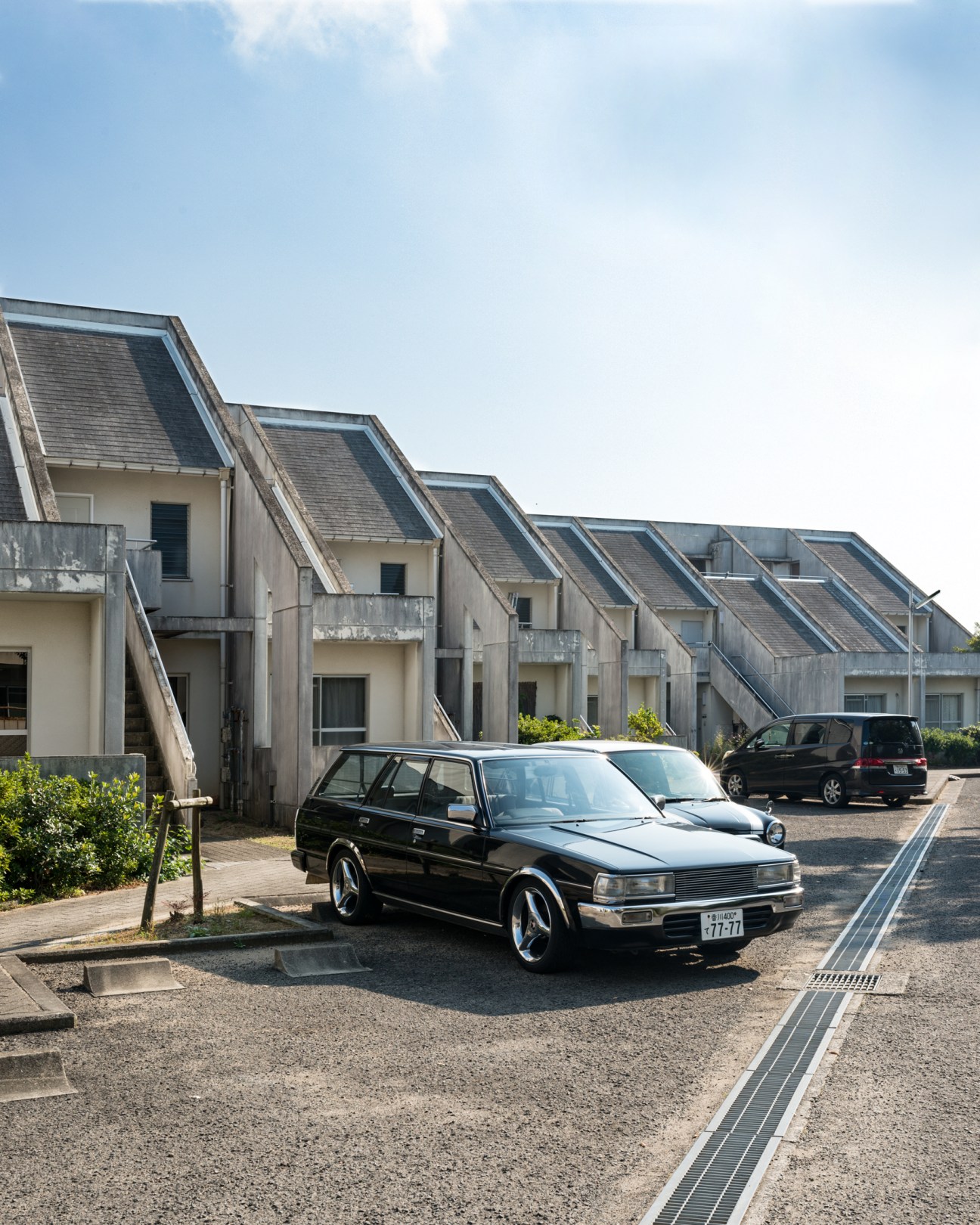 A black Nissan Bluebird estate car parked in front of modern residential houses with distinctive angled roofs