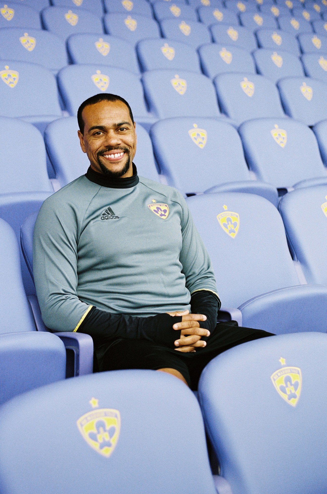 Marcos Tavares, Captain of NK Maribor FC, smiling while seated in blue stadium seats wearing team training gear