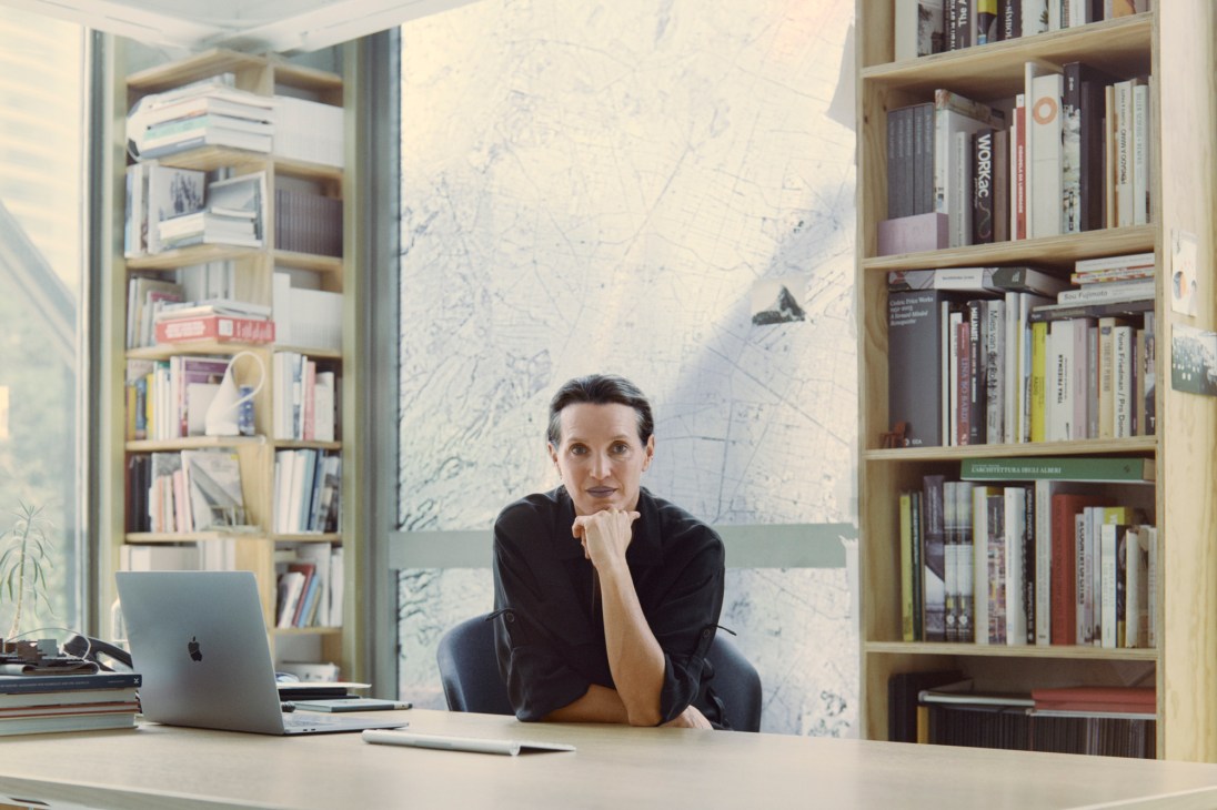 Tatiana Bilbao sits at a desk in her studio, surrounded by wooden bookshelves and architectural plans on the wall.