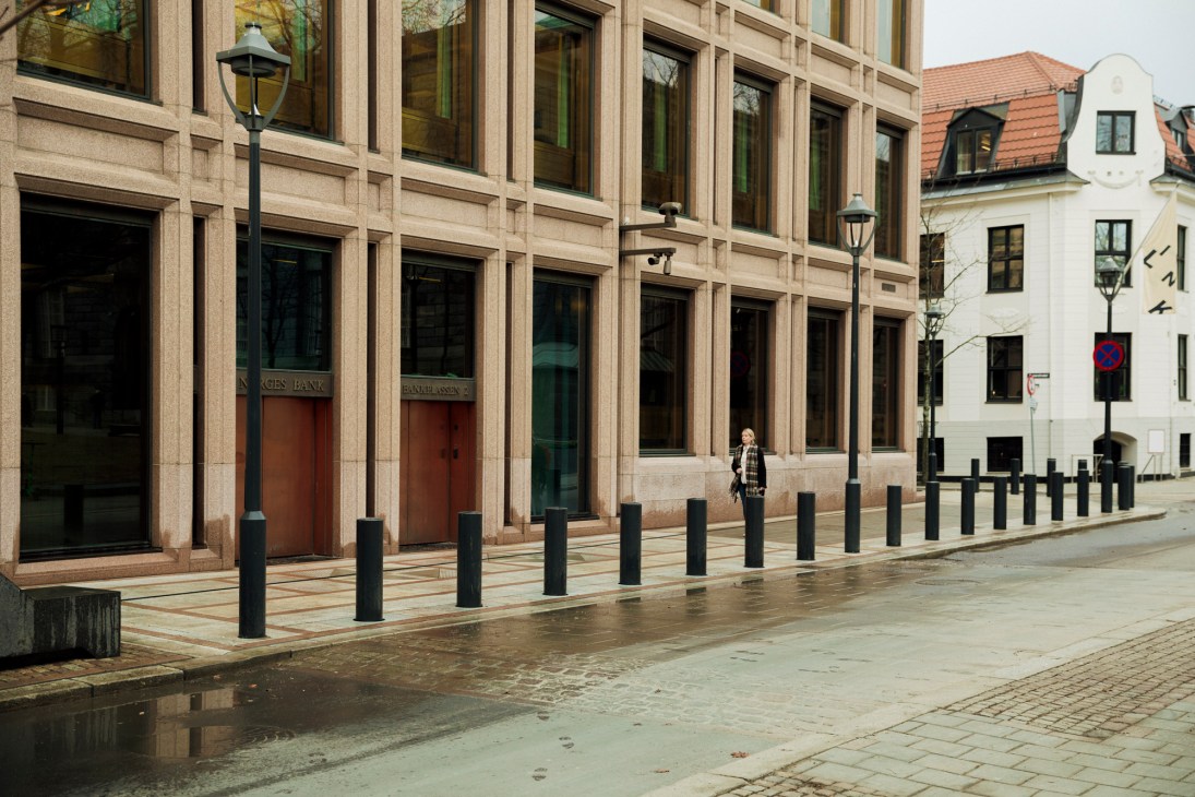 Stone building facade with large windows and bollards along wet cobblestone street in Oslo