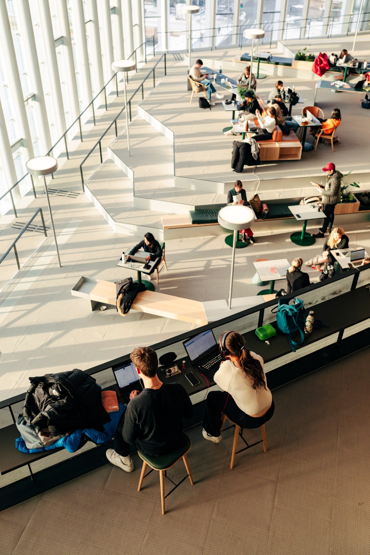 Students with laptops studying at tables on multiple levels of modern library interior with white stairs