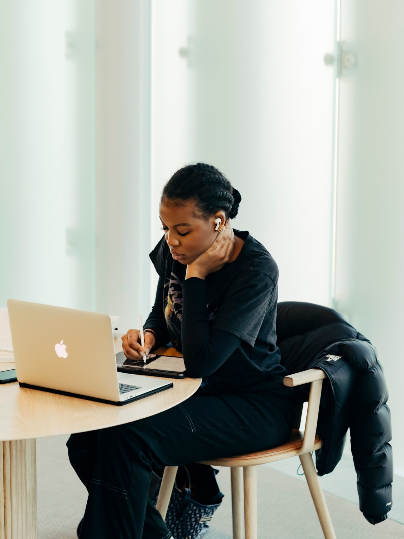 Person in black clothing working on laptop with drawing tablet at wooden desk by window