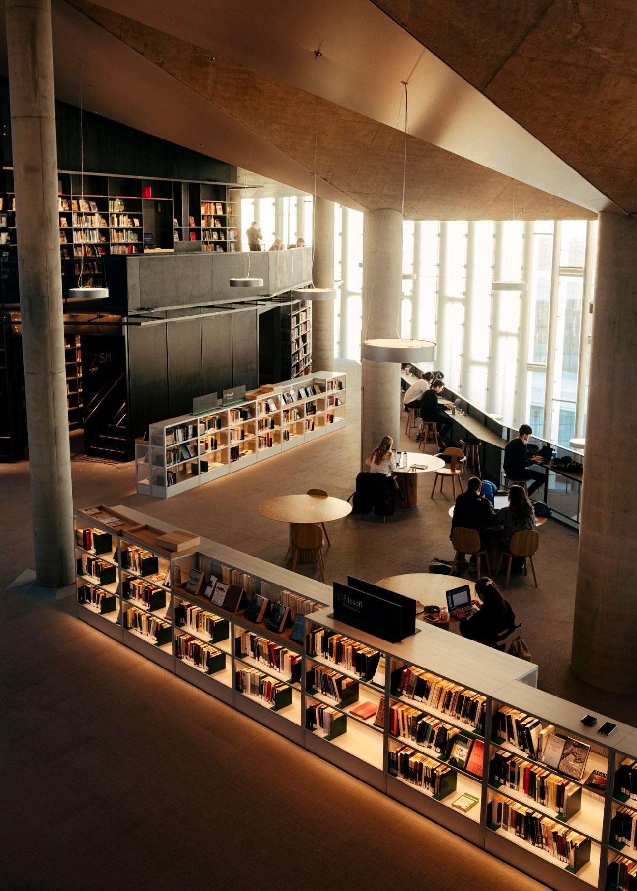Multi-level library interior with bookshelves, reading tables, large windows and people studying