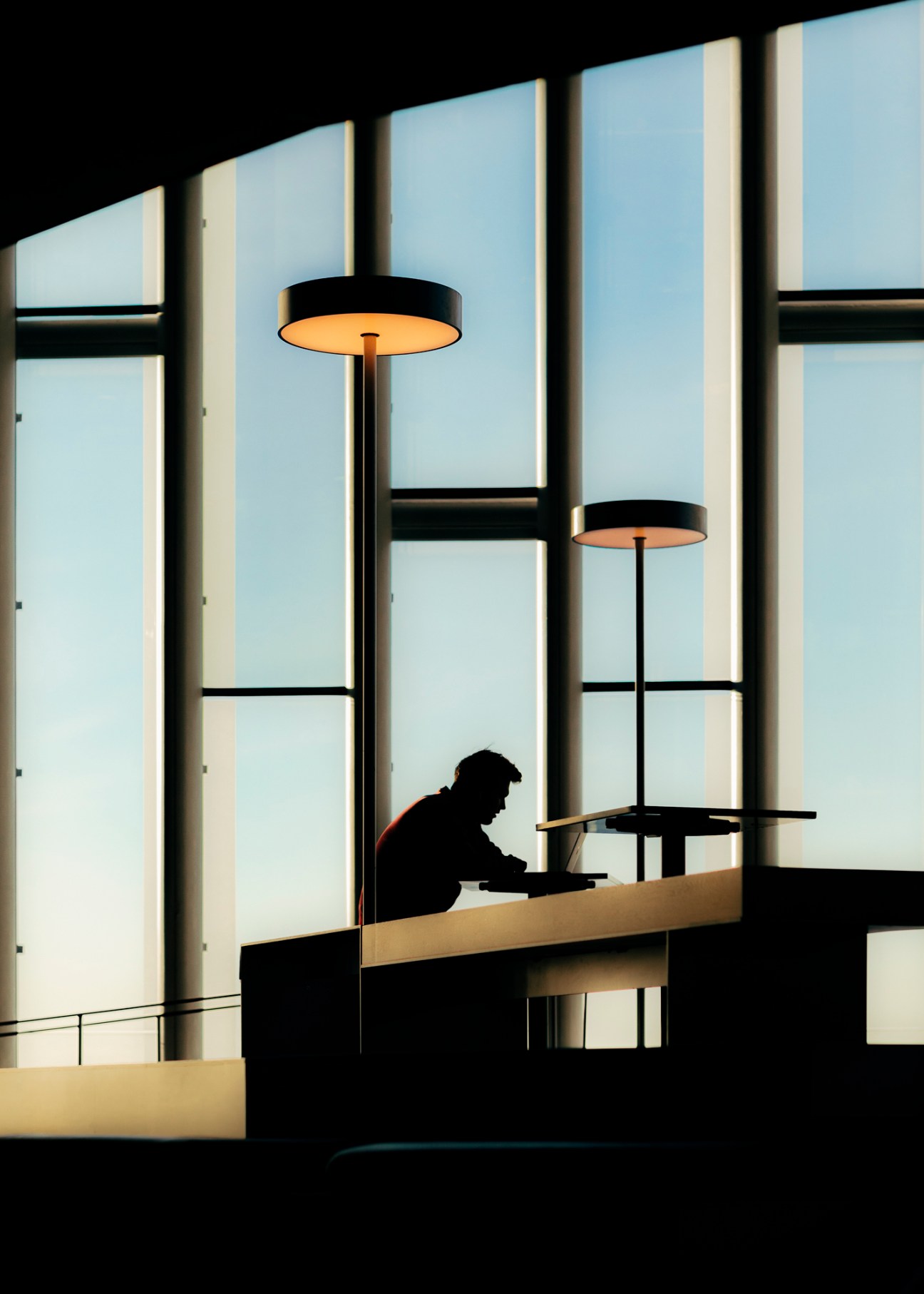 Silhouette of person reading at table by large windows in modern library interior with pendant lighting