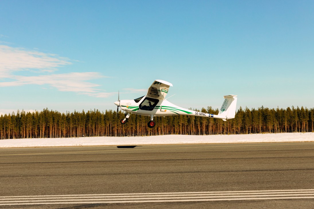 White electric aircraft taking off from runway with dense forest in background