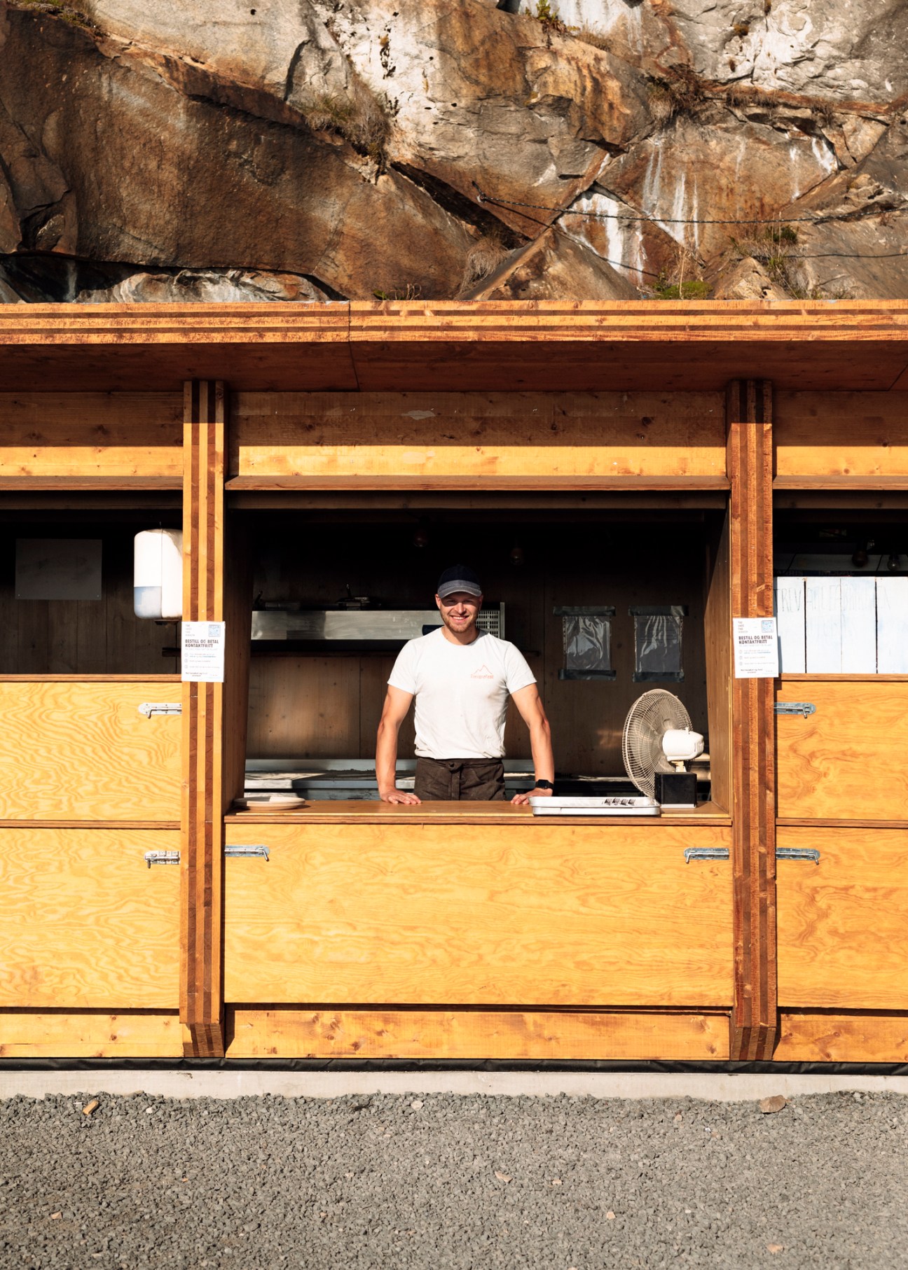 Man in white t-shirt and cap standing at wooden counter of pizza shop built into rock cliff.