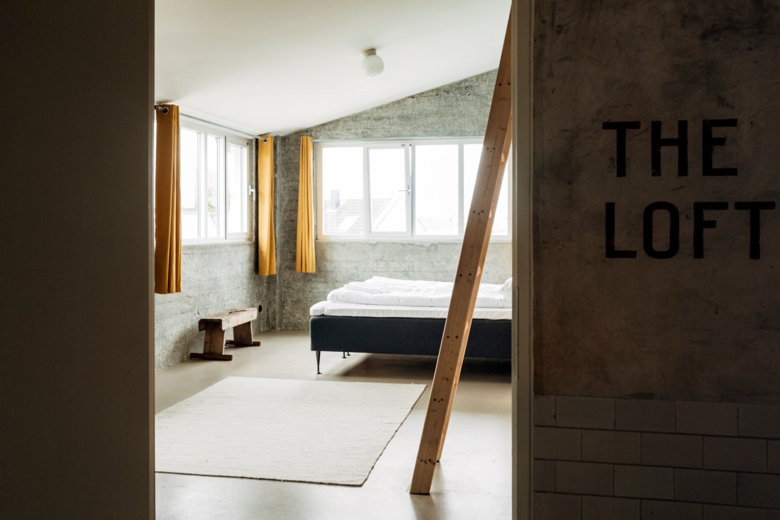 Modern loft bedroom with wooden beam, concrete walls, large windows, yellow curtains, and a bed with white linens.