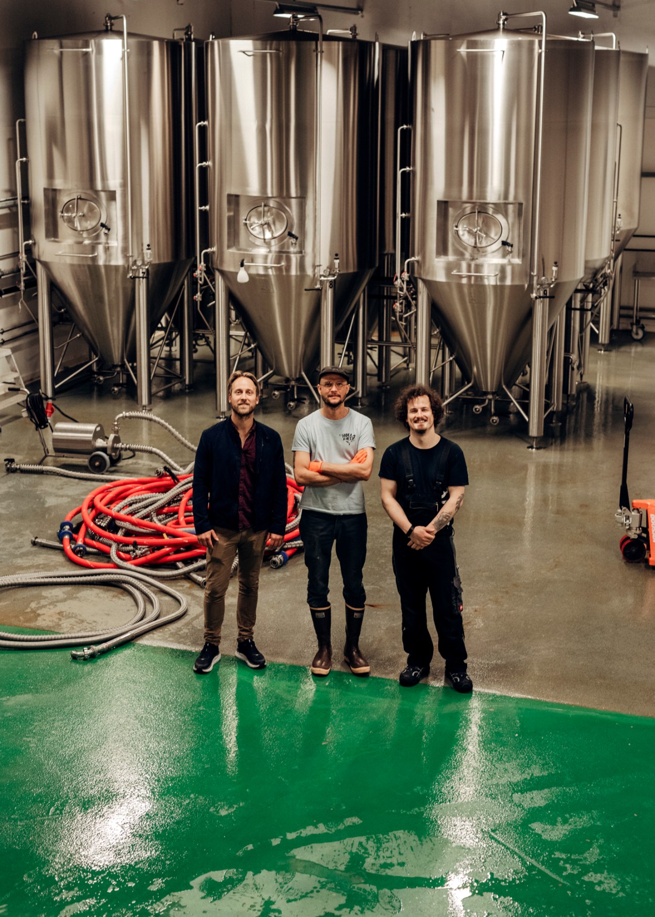 Three men stand in a brewery facility with large stainless steel fermentation tanks in the background and coiled hoses on the floor.