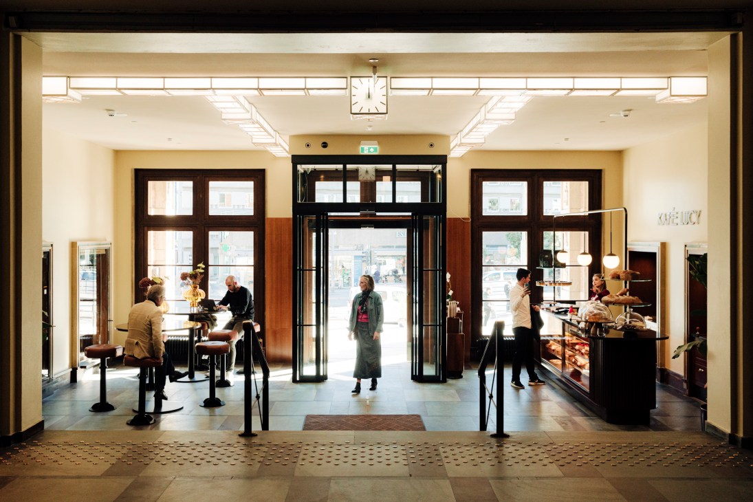 Hotel lobby entrance with glass doors, people sitting at tables and browsing displays, clock overhead